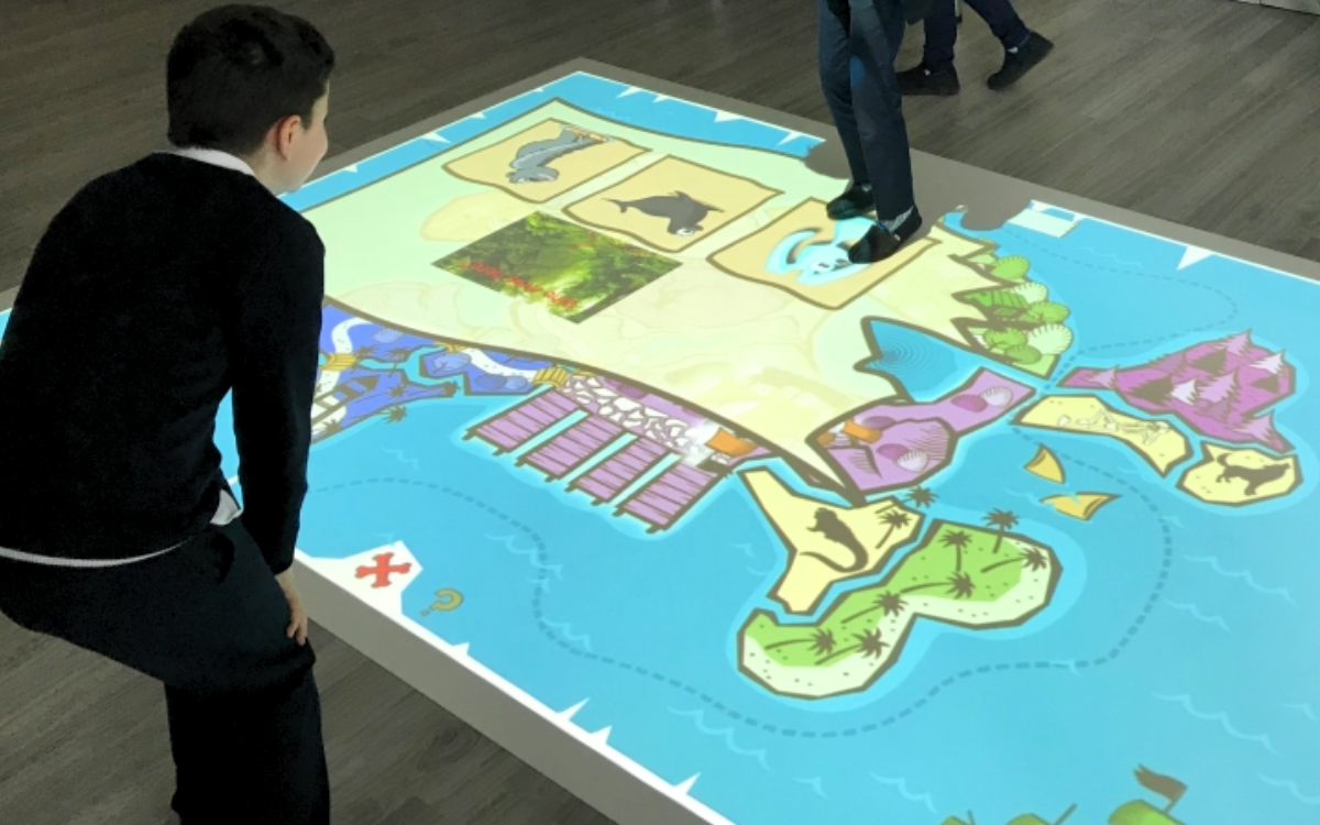 School children interact with each other as they participate in a dynamic learning experience with an ActiveFloor mobile projector, with vibrant images and educational content projected onto the floor. 